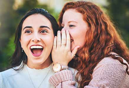 A young person whispering into her friends ear and she is smiling.